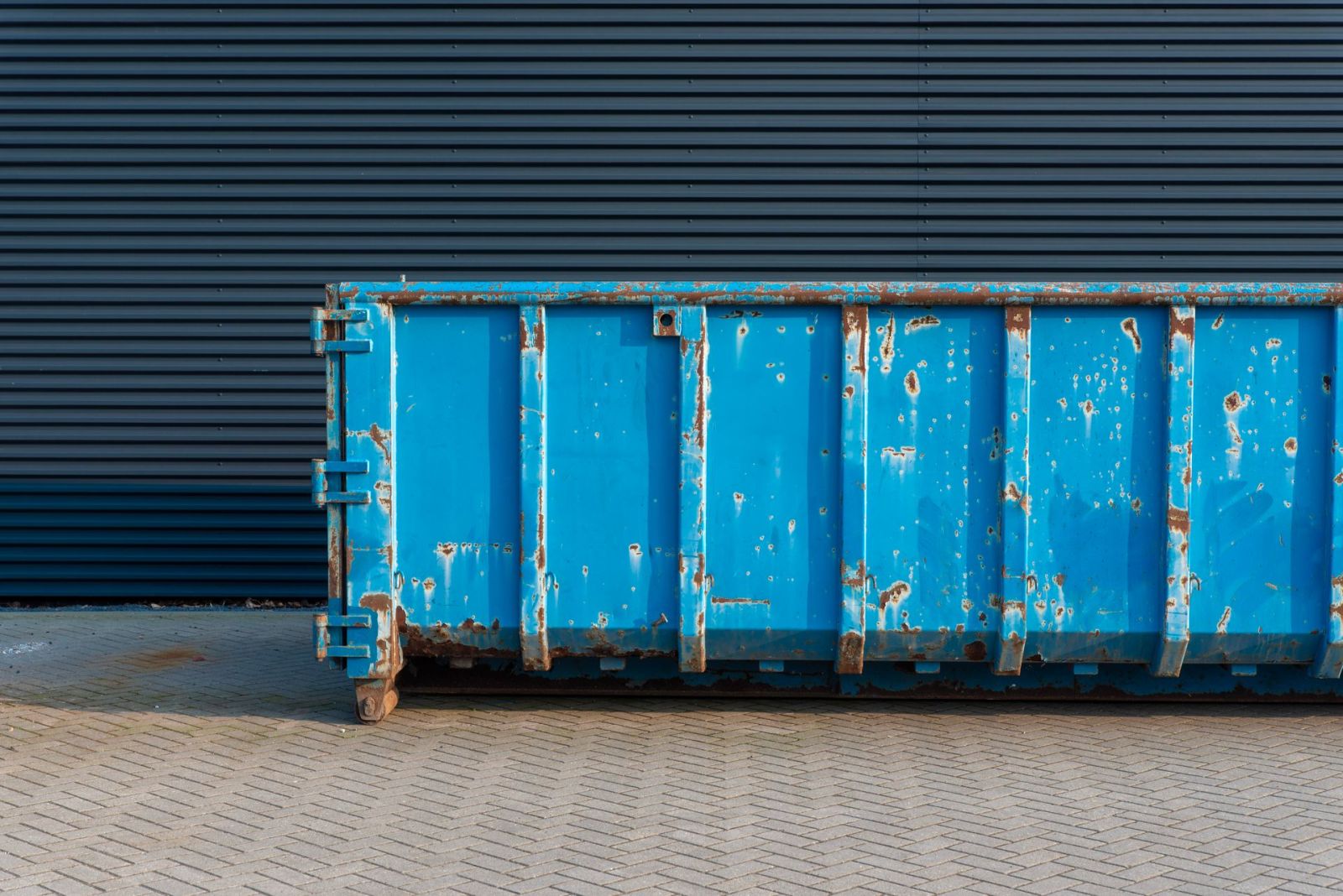 A large blue roll-on roll-off skip container standing on a paved commercial yard beside a steel-clad warehouse.