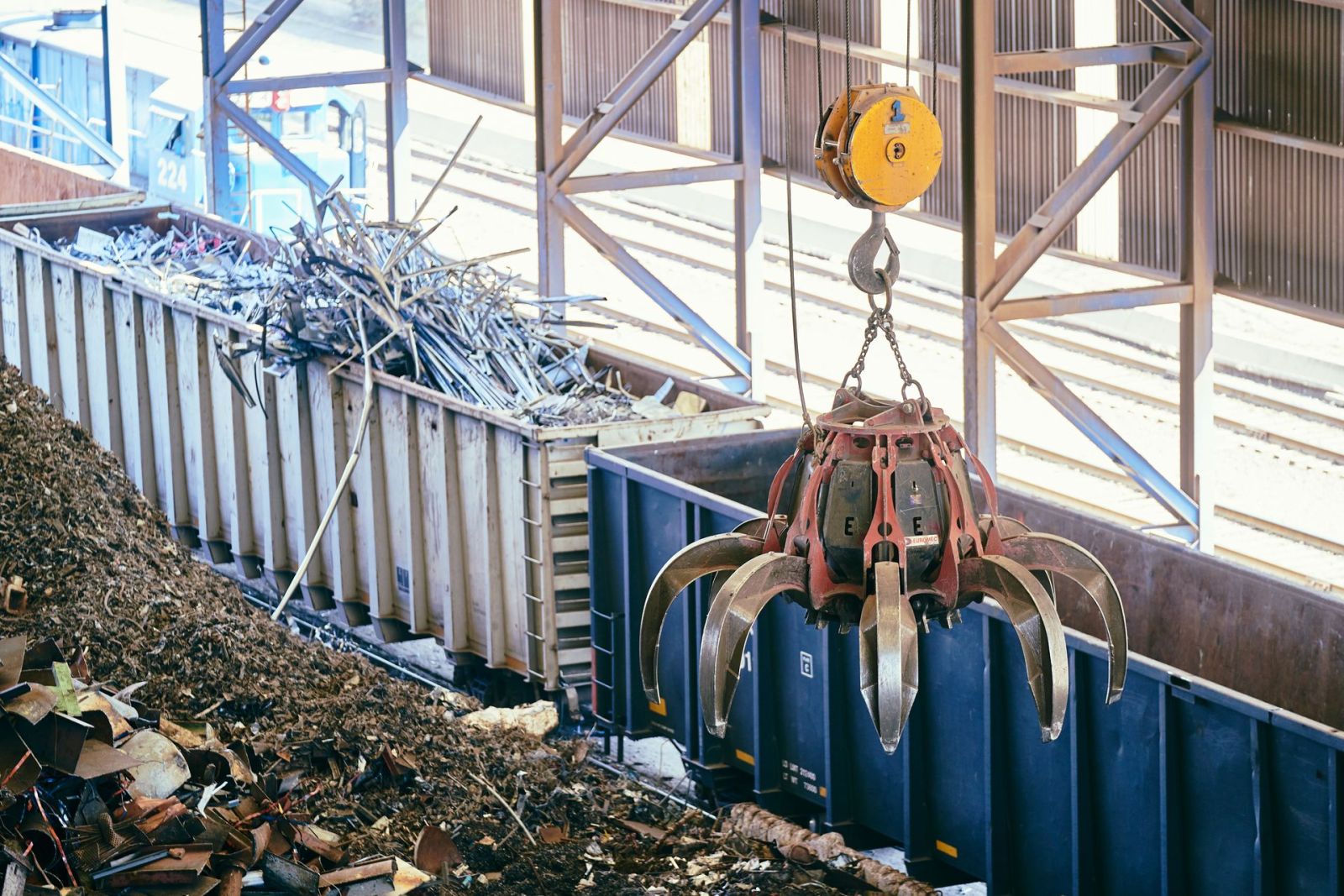 A materials-handling grab crane loading scrap metal waste into an open container at a licensed waste facility.